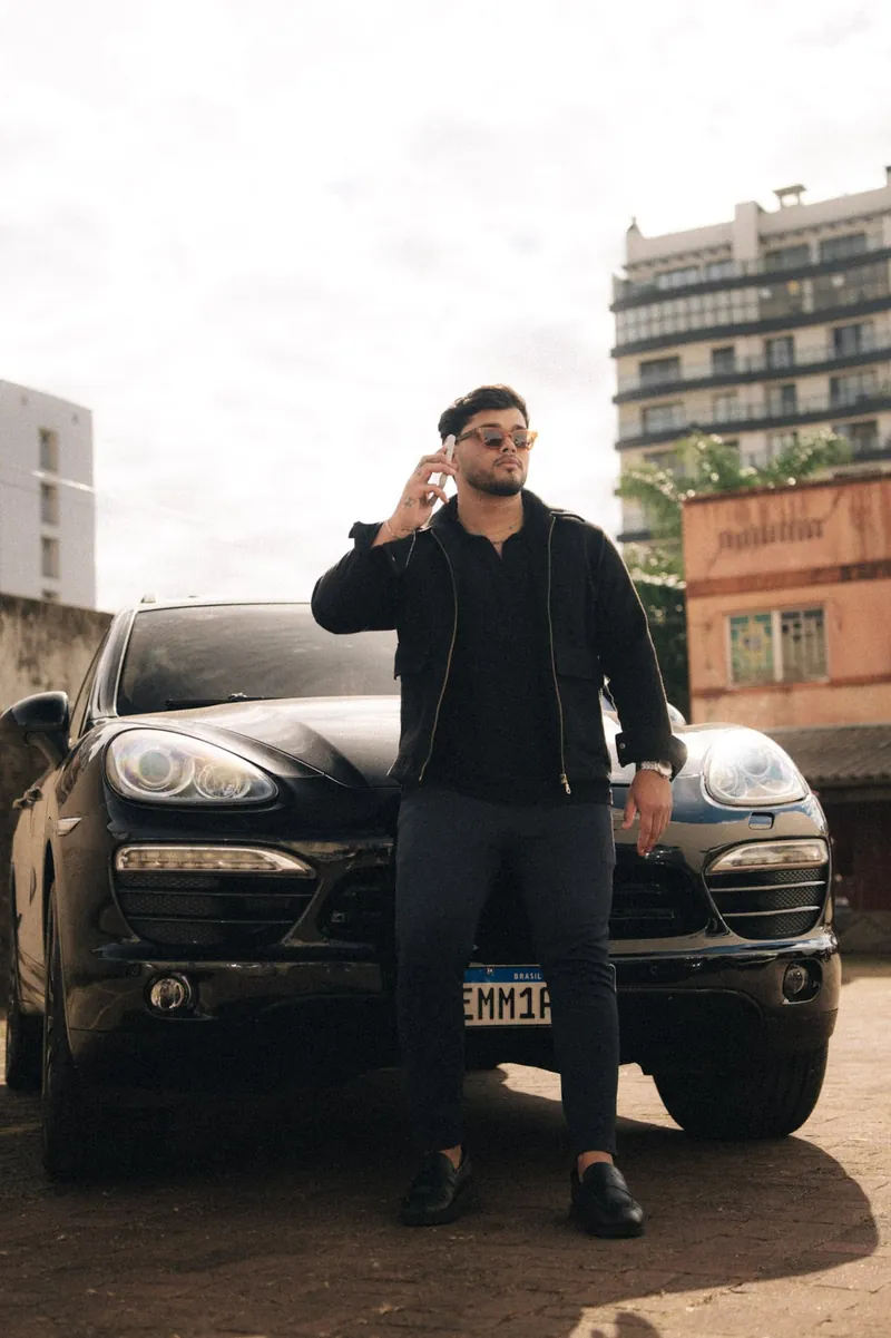 Aluizio Batu em pé ao lado de veículo, look em preto